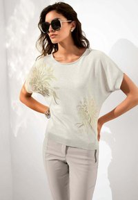 Lichtgrijze sweater met korte mouwen en bloemenborduurwerk, gecombineerd met lichtgrijze getailleerde broek en oversized zwarte zonnebril. Soepele textuur.