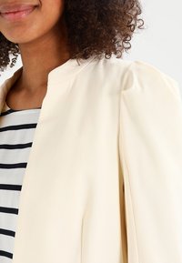 Blazer beige chiaro con una vestibilità rilassata. Presenta un colletto rotondo e un tessuto morbido. Indossato sopra un top a righe bianche e nere.