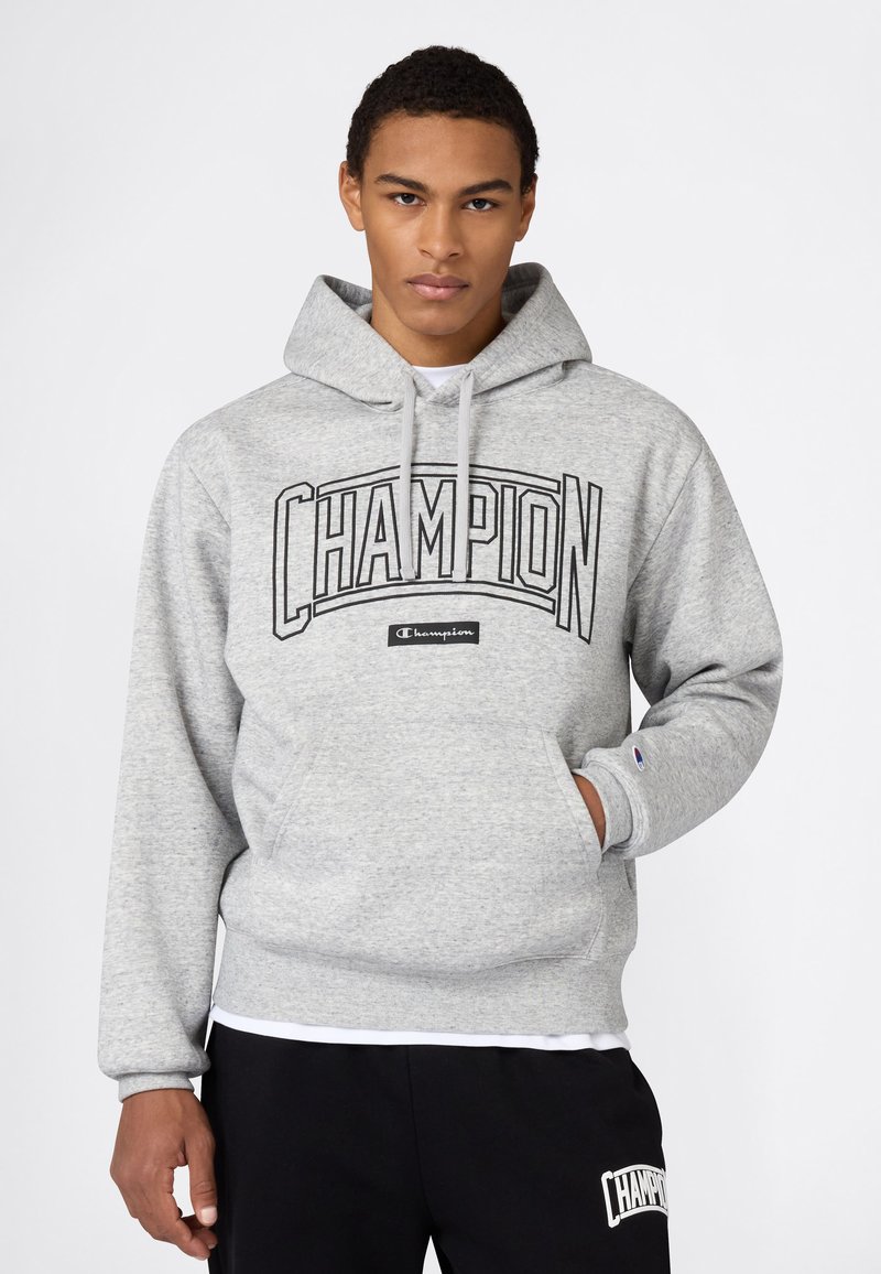 Sweat-shirt gris à capuche, grand logo "CHAMPION" en noir, poche kangourou, poignets et ourlet côtelés. Porté avec un pantalon noir.