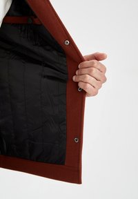 Röd ull ytterlager av en jacka, uppknäppt för att avslöja en slät svart foder med sömmar och interna fickor. Hand som greppar kanten.