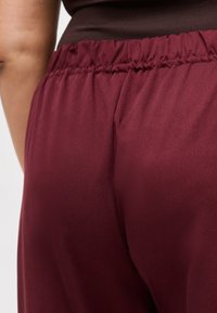 Κοντινό πλάνο ενός ατόμου που φοράει βερμούδες με ελαστική μέση σε μαρόν χρώμα και σκούρο μπλουζάκι, εστιάζοντας στη μέση και την υφή του υφάσματος.