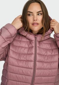 Kvinne som justerer hetten på en lilla vattert dunjakke, ser litt mot høyre mot en ensfarget bakgrunn.