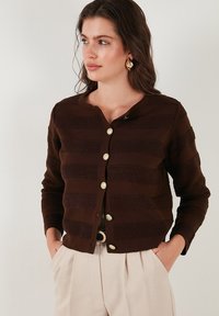 Cardigan en tricot marron avec des rayures texturées, col rond et boutons dorés. Porté sur un pantalon taille haute couleur crème.