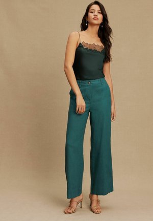 Satin dark green camisole with lace trim, paired with high-waisted turquoise wide-leg trousers. Beige strappy heels complete the outfit.