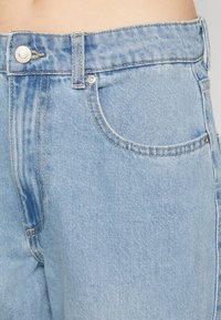 Jeans de mezclilla de color azul claro con cintura alta, costuras visibles, un bolsillo delantero y un acento de botón metálico en la cintura.