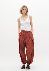 Hnedé plisované nohavice s širokými nohavicami a bočnými vreckami, skombinované s bielym priliehavým tielkom. Čierne šľapky dokončujú outfit.