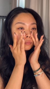 En person med långt, svart, vågigt hår använder sina fingrar för att applicera en produkt under ögonen, vilket visar välskötta naglar med ett mönster.