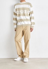 Pull rayé crème et beige avec le logo "HUGO", associé à un pantalon beige et des baskets blanches. Matière en tricot doux et poignets côtelés.