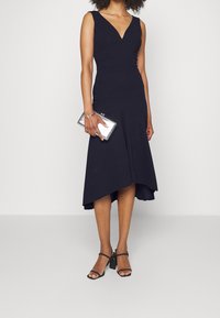 Abito senza maniche di colore navy con scollatura a V, corpetto aderente e orlo svasato. Il modello tiene una pochette argentata e indossa sandali neri con tacco.