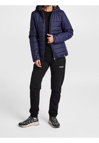 Chaqueta de plumas azul marino con capucha, diseño acolchado vertical, bolsillos con cremallera. Combinada con pantalones negros y zapatillas deportivas grises con acentos.