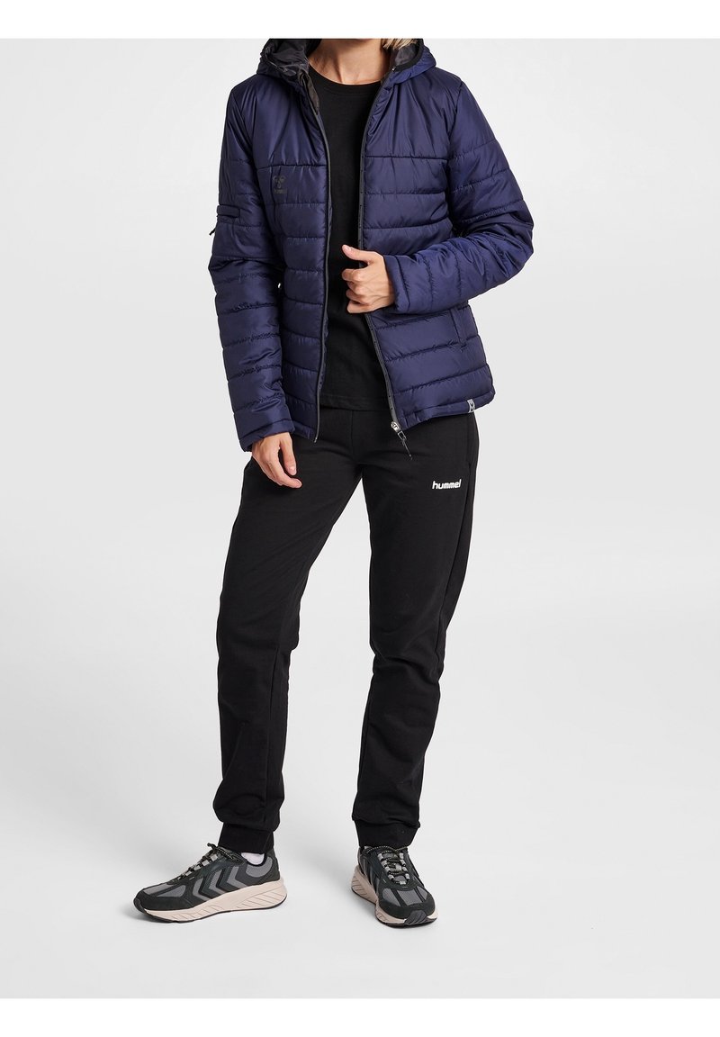 Chaqueta de plumas azul marino con capucha, diseño acolchado vertical, bolsillos con cremallera. Combinada con pantalones negros y zapatillas deportivas grises con acentos.