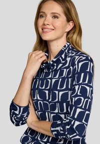 Junge Frau lächelt, trägt ein langärmliges Oberteil mit Marineblau-weißer Musterung und Wasserfallausschnitt, hält mit einer Hand den Ausschnitt.