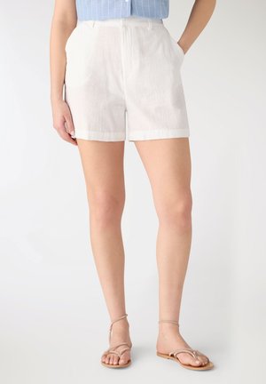 Donna che indossa pantaloncini bianchi su misura, camicia a righe blu infilata e sandali piatti color nude, in piedi con una mano in tasca.