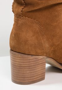 Bruna mocka ankelskor med en något rundad tå, texturerad yta och klack av staplad trä, med sömnads- och konturdetaljer.