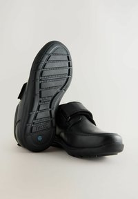 Des chaussures en cuir noir avec une sangle Velcro, un bout rond, et une semelle en caoutchouc texturée avec des rainures parallèles et le logo de la marque sur la semelle intérieure.