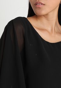 Svart genomskinlig topp med en bred halsringning, flödande ärmar och en mjuk textur. Anmärkningsv ärda detaljer inkluderar små imperfektioner i tyget.