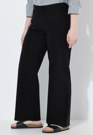 Person trägt schwarze Hosen mit weitem Bein, ein gestreiftes langärmliges Shirt und schwarze Slip-On-Sandalen mit Korksohle und steht an einer weißen Wand.