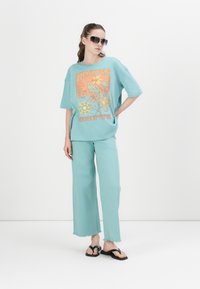 Camiseta turquesa de gran tamaño con diseño gráfico floral, combinada con pantalones anchos a juego. Chanclas negras y gafas de sol grandes completan el atuendo.