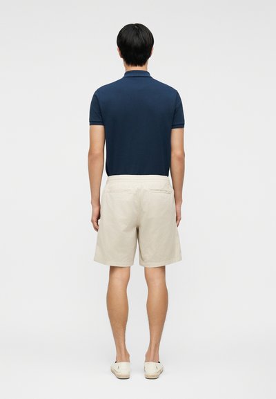 Άνδρας που στέκεται πλάτη, φοράει μπλε ναυτικό πόλο με κοντά μανίκια, μπεζ σορτς και ανοιχτόχρωμα παπούτσια χωρίς κορδόνια σε λευκό φόντο.