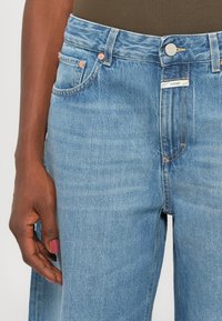 Main gauche avec vernis à ongles rose reposant à côté d'un jean en denim bleu clair avec un bouton en métal et une petite étiquette blanche portant l'inscription "CLOSED."