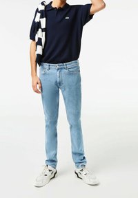 Polo bleu marine à manches courtes, emblème vert, jean slim bleu clair, et baskets blanches avec des accents noirs. Pull drapé sur les épaules.