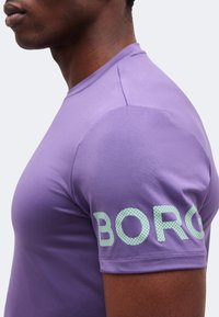 Άνδρας που φοράει εφαρμοστό μωβ T-shirt με ανοιχτοπράσινα ανάγλυφα γράμματα "BORG" στο μανίκι, εμφανίζεται από το πλάι σε απλό φόντο.