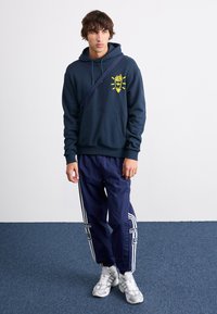 Hanorac bleumarin cu grafic galben, asortat cu pantaloni de trening bleumarin cu dungi albe și brand. Completat cu adidași gri.