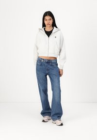 Gray cropped zip-up hoodie with a front pocket, paired with blue high-waisted wide-leg jeans and light sneakers. Simple, casual look.
