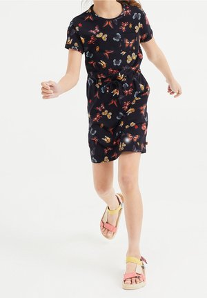 Giovane con un vestito nero a maniche corte con motivi colorati di farfalle e insetti che corre, indossando sandali open-toe con cinturini rosa e gialli.