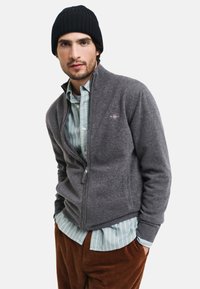 Grå zip-up cardigan gjord av ett mjukt stickat material, bärs över en ljusblå och vit randig skjorta, tillsammans med bruna kordflöjlsbyxor. Svart mössa.