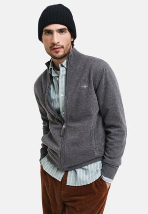 Cárdigan gris con cremallera hecho de un material de punto suave, usado sobre una camisa a rayas azul claro y blanca, combinado con pantalones de pana marrones. Gorro negro.