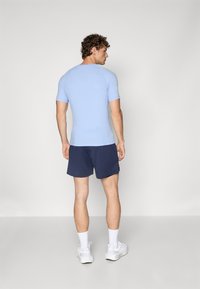 Männliches Sportoutfit mit einem hellblauen, enganliegenden Kurzarmhemd und dunkelblauen Shorts aus leichtem Stoff, kombiniert mit weißen Sneakers.