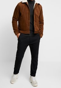Veste en velours côtelé marron avec un col en fausse fourrure blanche, associée à un pull foncé et un pantalon noir. Les chaussures sont des baskets de couleur claire.