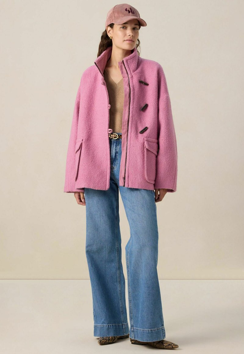 Femme portant une veste en polaire rose surdimensionnée, un pull beige clair, un jean bleu à jambes larges, des chaussures en serpent et une casquette de baseball rose.