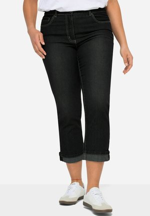 Pantalones capri de mezclilla negra con dobladillo enrollado, cinco bolsillos y costuras de contraste, combinados con zapatillas blancas.