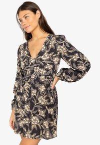Vestido mini floral en negro con flores en crema y dorado. Presenta un escote en V, mangas largas y una cintura fruncida. Tejido suave y texturizado.