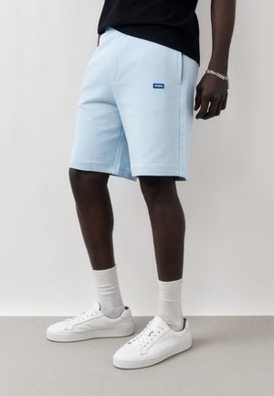 Hombre que lleva pantalones cortos Hugo azul claro, calcetines blancos y zapatillas blancas, de pie sobre un suelo gris con un fondo sencillo.