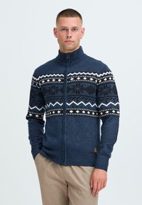 Jūrinės spalvos zip-up megztinis su grublėta apykakle ir rankogaliais. Turi juodai baltą raštuotą dizainą per krūtinę, pagamintas iš tekstūruoto audinio.