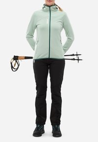 Veste zippée vert pâle avec un col haut, associée à un pantalon noir. La tenue comprend des bâtons de trekking à l'arrière et des chaussures de randonnée noires avec des accents turquoise.