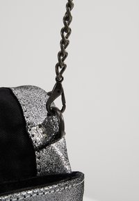 Tracolla a catena in metallo collegata a una borsa metallica argentata, lucida e strutturata. La borsa presenta dettagli in tessuto nero e un bordo liscio.