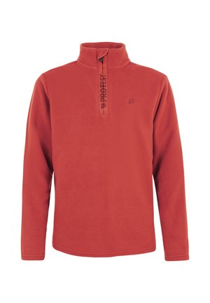 Niño sonriente con pelo rizado que lleva un forro polar rojo con cremallera hasta el pecho, pantalones verdes de exterior y zapatos negros, de pie sobre un fondo blanco.