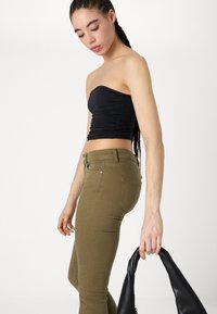 Top negro sin tirantes con detalles fruncidos, combinado con pantalones ajustados de color verde oliva. La modelo sostiene un bolso negro. Texturas suaves y diseño ajustado.
