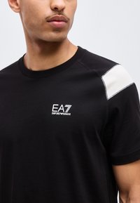 Νεαρός άνδρας που φοράει μαύρο μπλουζάκι EA7 Emporio Armani με λευκή επιγραφή στον δεξί ώμο, κοιτάζοντας ελαφρώς προς τα αριστερά.