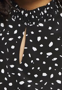 Svart blus med vita abstrakta fläckar. Har en smockad halsringning och en nyckelhålsöppning framtill. Slät tygtextur.