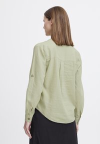Chemise verte clair à manches longues en tissu doux. Caractéristiques des accents de boutons sur les manches et un détail de pli dans le dos. Ourlet arrondi.