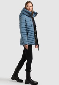 Giacca a vento azzurro chiaro con cuciture orizzontali, colletto alto, tasche con zip nere e una vestibilità leggermente rilassata. Indossata con leggins neri e stivali.