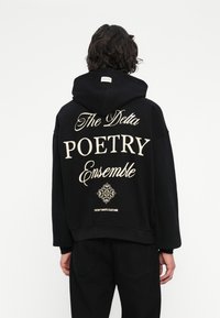Cilvēks ar tumšiem viļņainiem matiem, tērpies melnā kapučjakā ar uzrakstu "The Delta POETRY Ensemble" un dekoratīvu dizainu aizmugurē, stāv pret baltu fona.