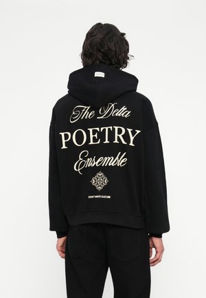 Osoba z ciemnymi falującymi włosami nosi czarną bluzę z kapturem z napisem "The Delta POETRY Ensemble" oraz dekoracyjnym wzorem na plecach, stojąc na tle białym.