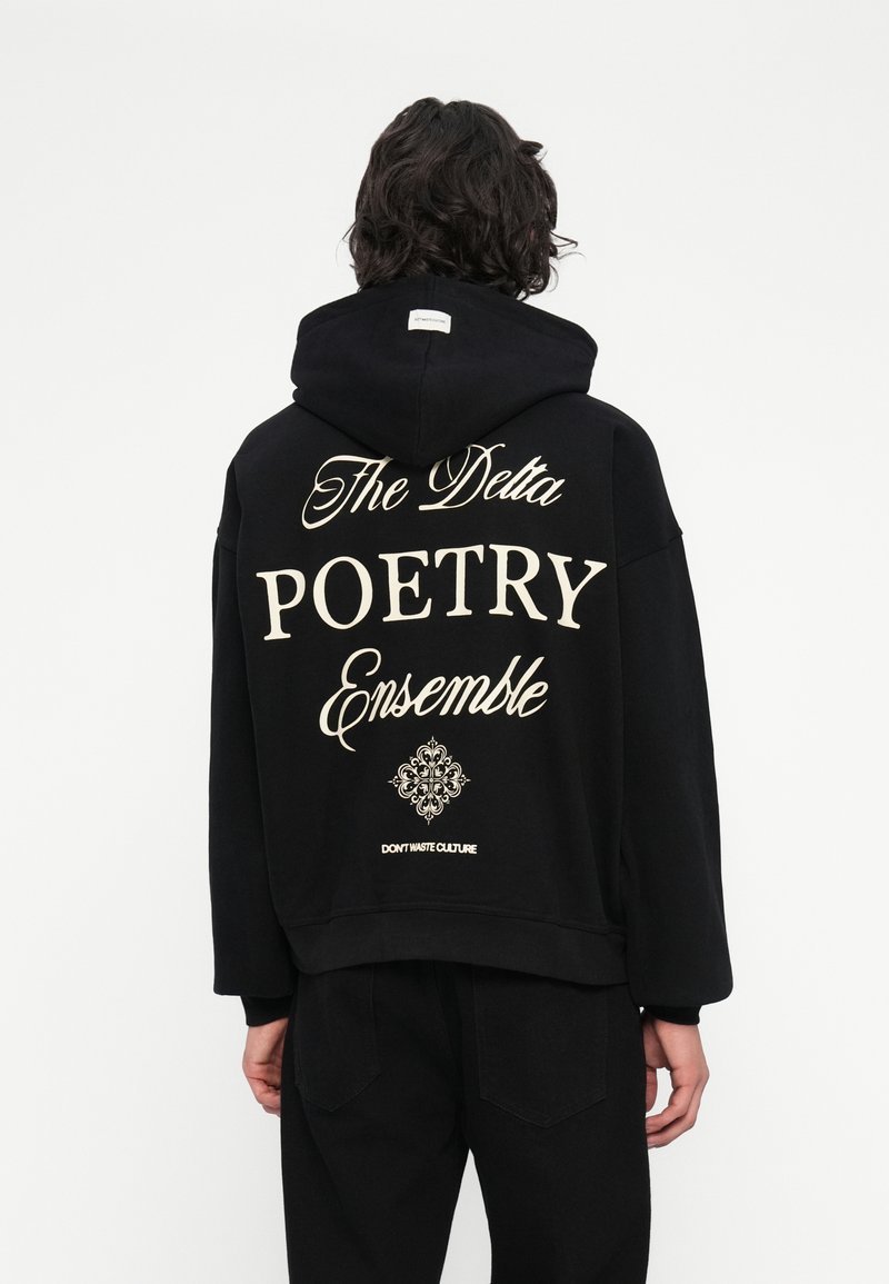 Cilvēks ar tumšiem viļņainiem matiem, tērpies melnā kapučjakā ar uzrakstu "The Delta POETRY Ensemble" un dekoratīvu dizainu aizmugurē, stāv pret baltu fona.