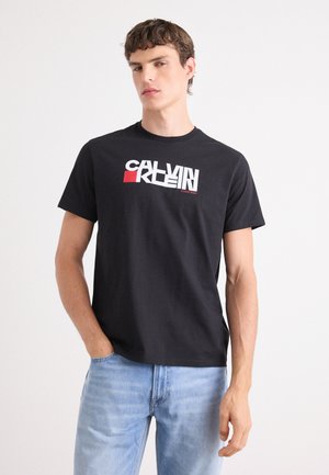 Black cotton t-shirt featuring a bold white and red "Calvin Klein" logo. Classic crew neck design with short sleeves and a relaxed fit.